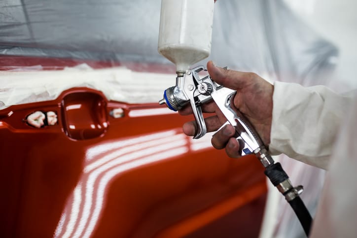 Spray gun laying fresh red paint across a masked car panel in the booth